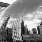 Black and white photo of a city skyline mirrored in a cloud gate, showcasing urban architecture and reflections.