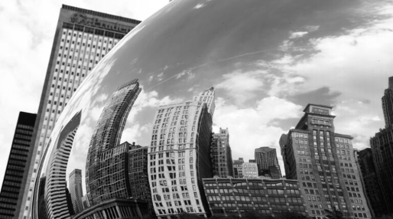 Black and white photo of a city skyline mirrored in a cloud gate, showcasing urban architecture and reflections.